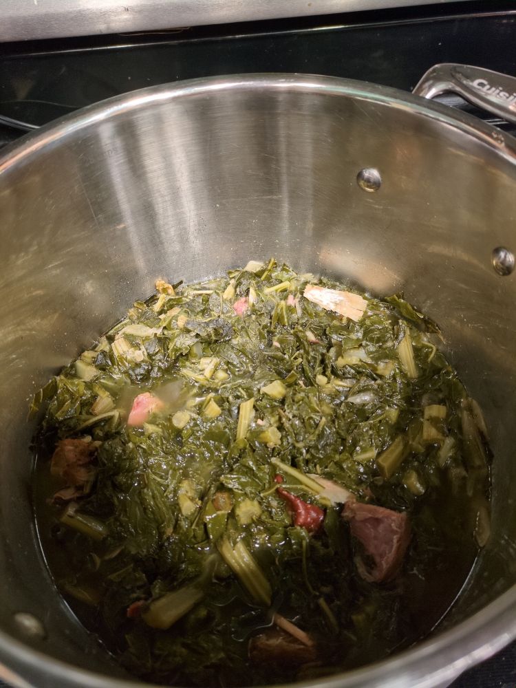 A tall rimmed steel pot with dark green, slow cooked collards simmering inside. Bits of smoked turkey can be seen amongst the greens in the murky pot liquor.