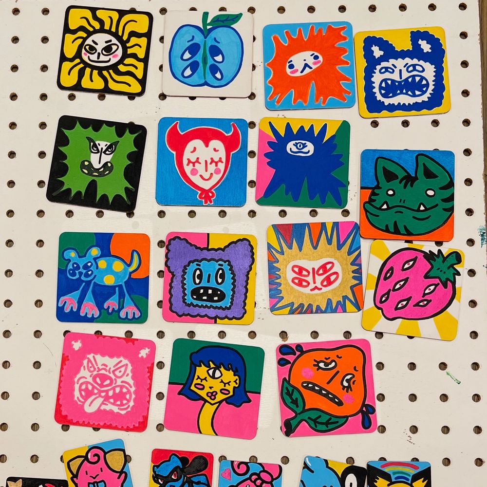 ID: a photo of 15 small painted wood pieces sitting next to each other on a pegboard. They all have bright colors and in order: a flower with a face, half an apple with four eyes, an orange spiky guy, angry blue cat kinda guy, green spiky guy, guy in a devil bonnet, a blue spiky guy, a green cat head with an underbite, a dog with weird large paws, purple and blue monster head, a gold spiky guy with rainbows surrounding himb, a strawberry with a bunch of eyes, an angry pink bear sticking his tongue out, a lady with three eyes and a long neck, and, a distressed peach.