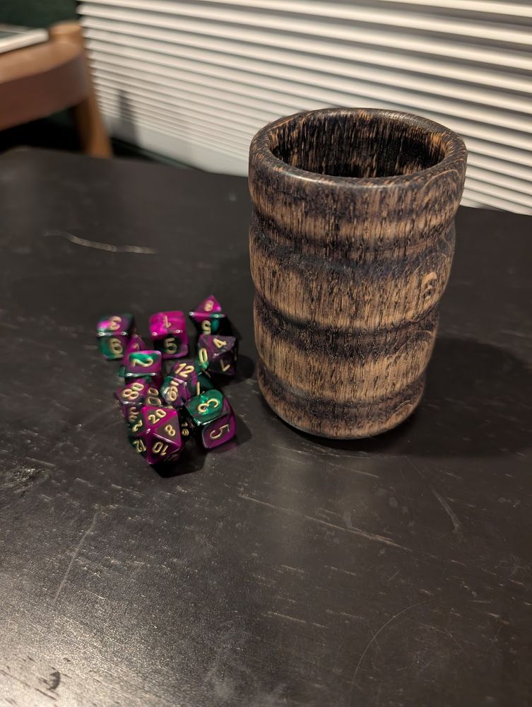 A wooden dice cup, made of ebonized red oak, standing upright beside a pile of dice