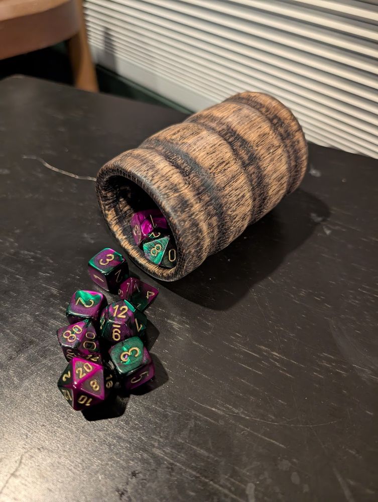 A wooden dice cup, made of ebonized red oak, laying on its side as dice spill out of it