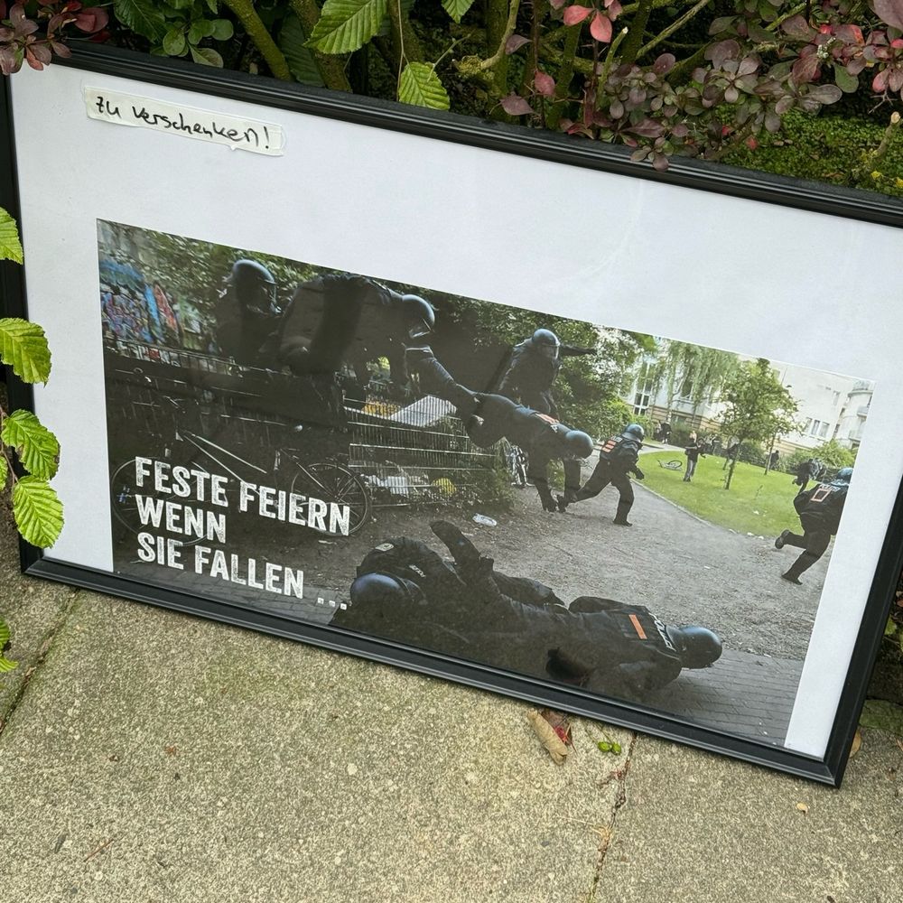 Ein Bilderrahmen der auf dem Boden steht und an einer Hecke angelehnt ist. Auf dem Bild sieht man Bullen die über einen Zaun springen. Ein paar liegen am Boden. Oben am Rand des Bildes steht „zu verschenken!“