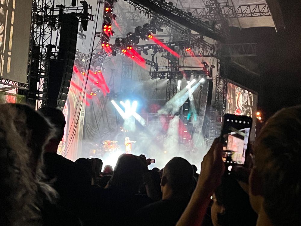 An image of a concert performed by the band Ghost. In this image you can sort of make out their lead singer wearing a red jacket under white and red stage lights. 