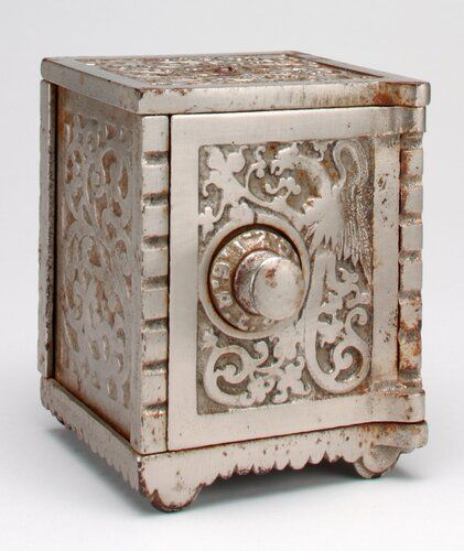 square silver bank safe with numeric combination knob at left center on door; "saving bank" at top surrounded by raised vine design; scrollwork at sides; coin slot at top back panel; 12 large holes drilled in base to see contents