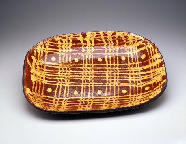 brown ground with yellow slip decoration on top of vertical and horizontal lines and 2 rows of dots
Redware decorated with slip patterns was an important part of the English and American folk tradition in ceramics during the 18th and 19th centuries. Light-colored slip (watered-down clay) was painted directly on the red clay body. Feathers and other tools were dragged through the slip to create animated patterns. A resurgence of interest in the slipware tradition occurred during the mid-20th century with ceramists such as the English potter Michael Cardew and the American Ken Ferguson.