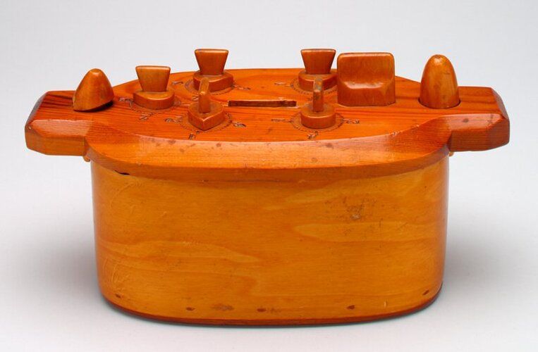 oval, wood; 5 dials and 3 other knobs on top; coin slot on top; braid (2 rows) on side; top rotates to open on peg next to 3 dials when combo is properly set