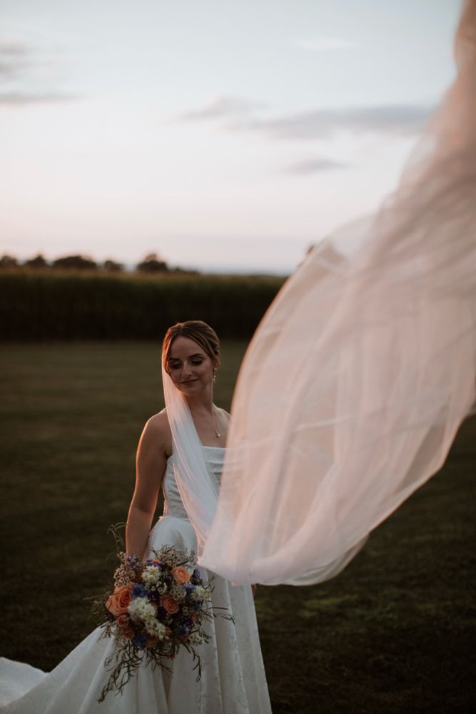 Cat on her wedding day, holding a big bouquet of wildflowers and her veil is flowing in the wind