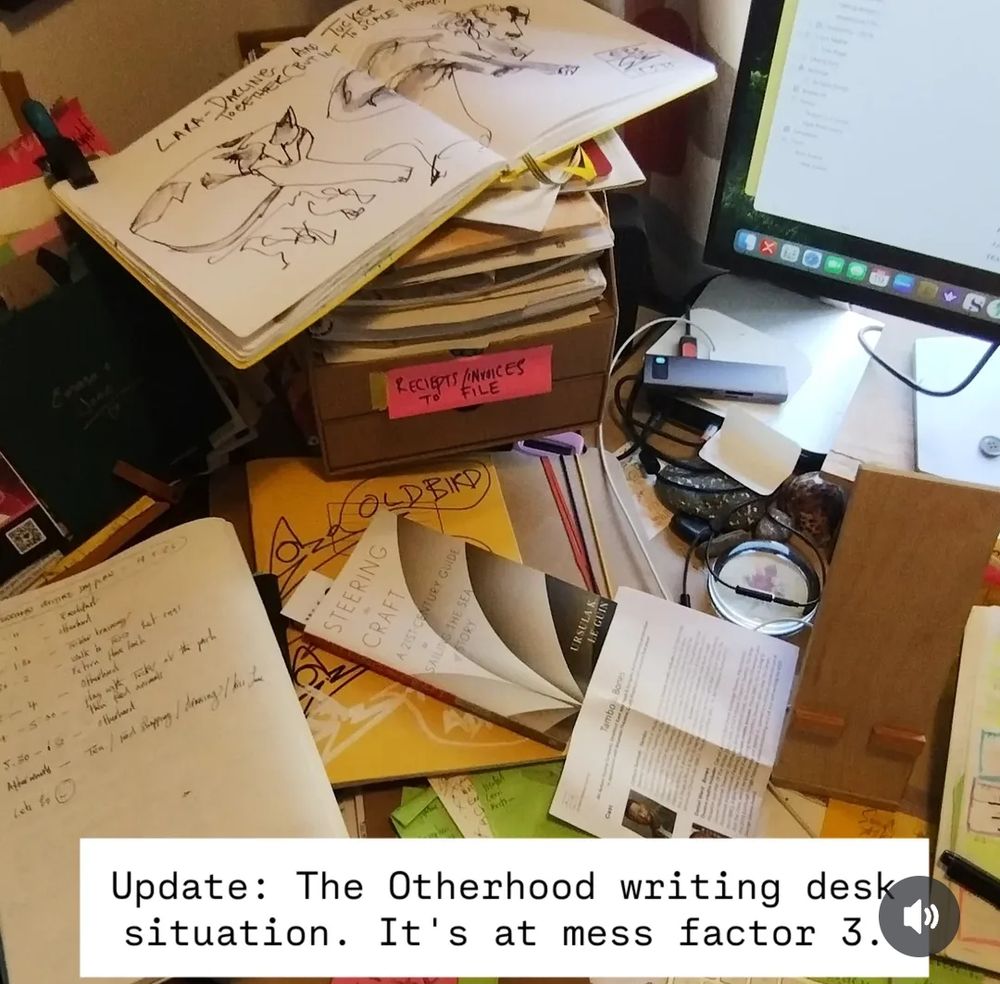 A mess of paper notebooks, a sketch book, paperweights a computer a book by Ursula K Le Guin 'Steering the craft', a theatre programme sheet. It's hideous. Text at the bottom reads, 'Update: The Otherhood writing desk situation. It's at mess factor 3.'