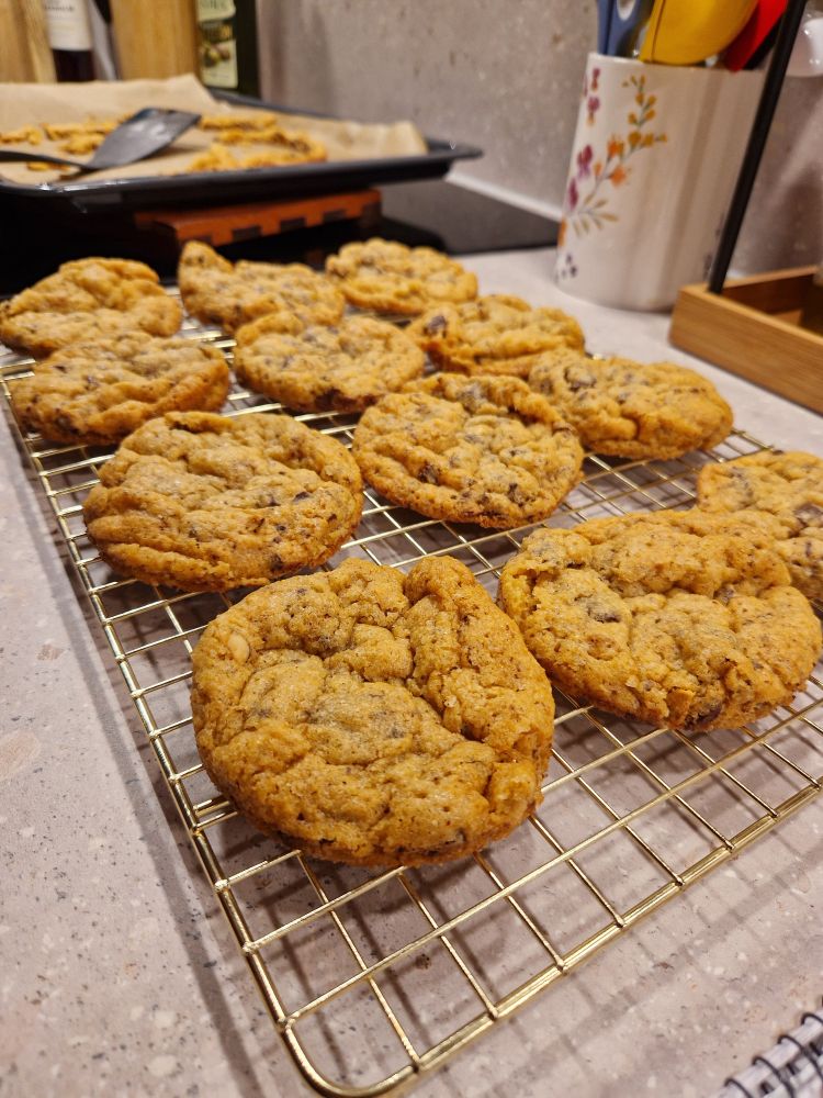 12 cookies med hakket chokolade og hasselnødder på en guld afkølingsrist.  I baggrunden kan man se rester fra kanterne på en bageplade