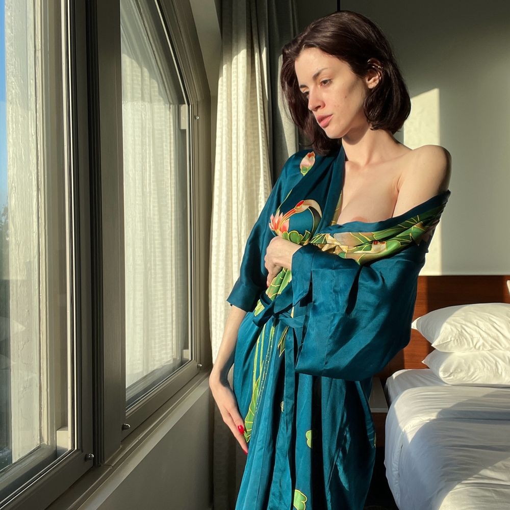 Lux Neon standing in a long silk robe that’s falling down her shoulder as she looks out a large window in the morning light