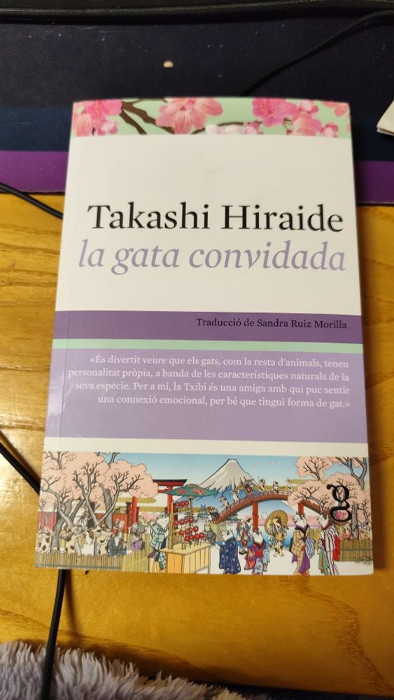 Foto de la portada del libro "La Gata Invitada" de Takashi Hiraide en catalán. 
El libro es (de arriba a abajo): una franja de flores de cerezo sobre fondo verde; blanco donde sale el nombre del autor y el título del libro; lila oscuro donde pone que lo ha traducido Sandra Ruiz Morilla; una pequeña franja verde pastel; una franja mediana lila pastel donde pone (en catalán) «Es divertido ver que los gatos, como el resto de animales, tienen personalidad propia, dejando de lado las características naturales de su especie. Para mí, Txibi es una amiga con quien puedo sentir una conexión emocional, por mucho que tenga forma de gato.»; luego una pequeña franja blanca; por último, una ilustración en estilo japonés de un festival tradicional de Japón donde los miembros del festival son gatos con ropajes tradicionales (kimonos, yukatas, etc).

El libro está encima de una mesa de madera. En la parte superior se ve parte de un tapete para teclado y ratón de color lila oscuro y negro. En la parte inferior, pasando por debajo de la esquina inferior izquierda del libro, hay un cable negro.

Por último, se ve parte de mi batín de andar por casa en la parte más inferior de la foto. Es peludito de color lila pastel.