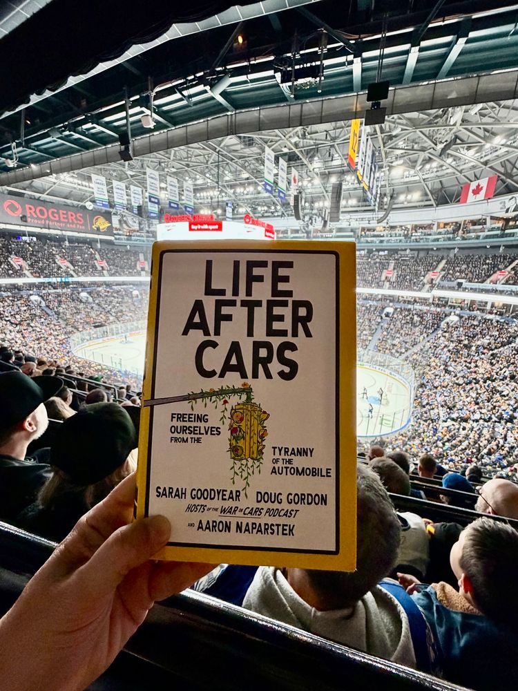 Life After Cars book cover held up in hockey arena wink rink in background. 