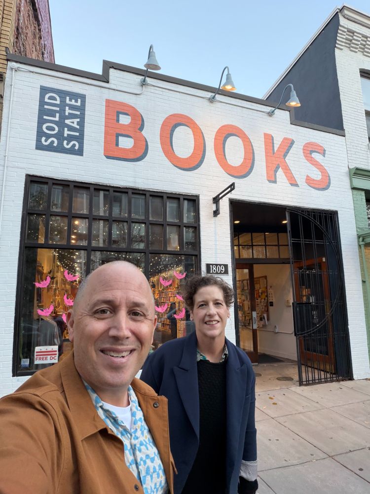 Doug Gordon in brown jacket and Sarah Goodyear in navy jacket in front of white storefront with "SOLID STATE BOOKS" painted across the top.