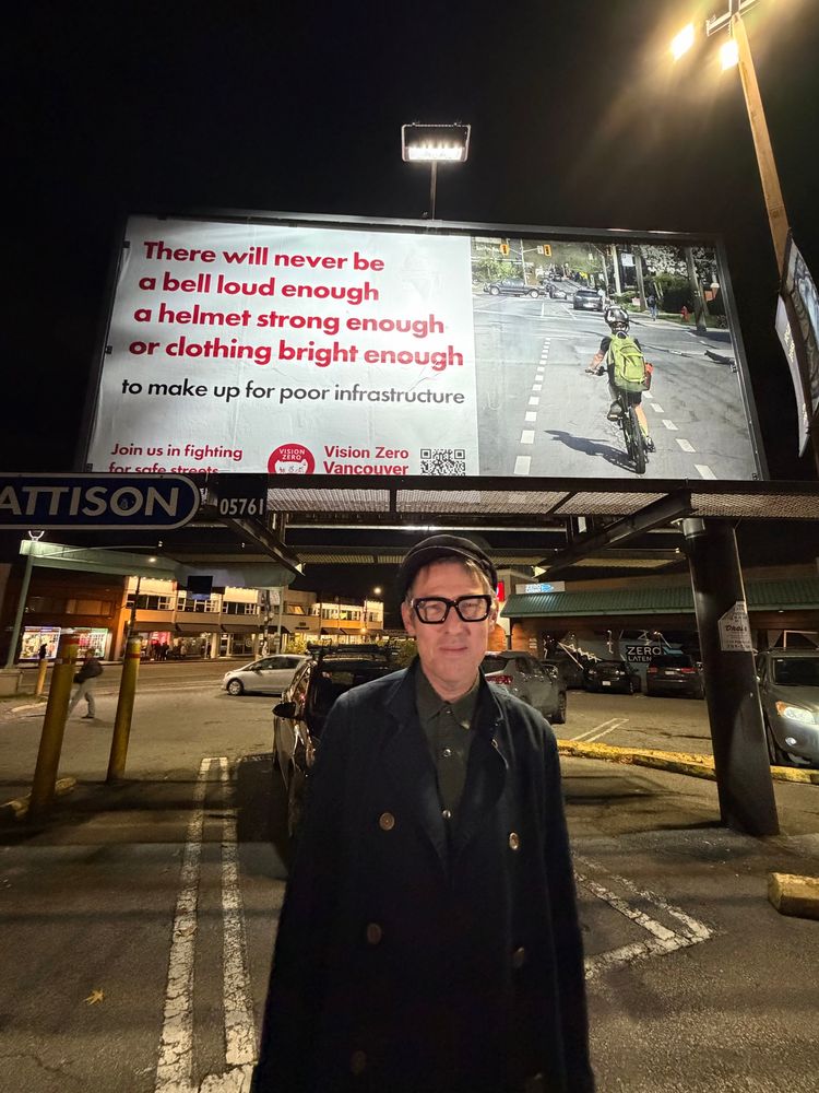 Tom Flood in black jacket and black hat in front of billboard that says 
There will never be a bell loud enough, a helmet, strong enough or clothing bright enough to make up for poor infrastructure. Billboard shows a picture of a child with a green backpack, riding a bicycle and a painted bicycle lane.