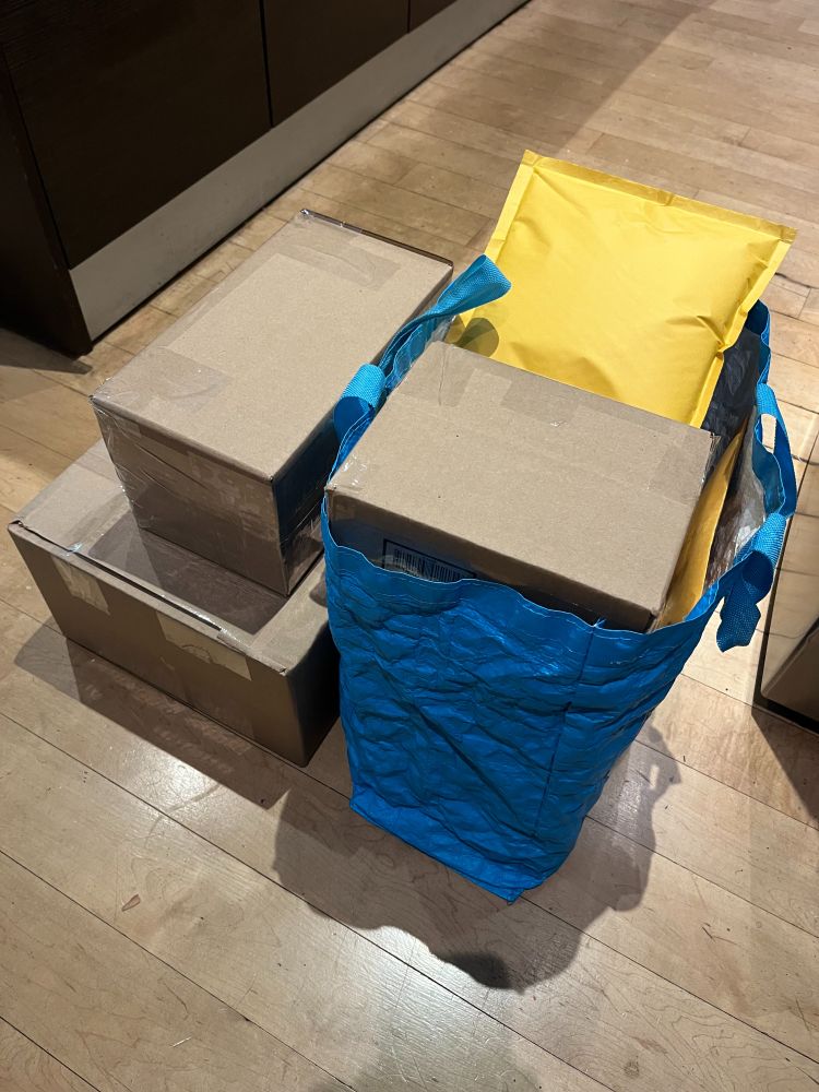 Cardboard boxes on light wood floor, some are in blue plastic bag. There is a yellow padded envelope on top.