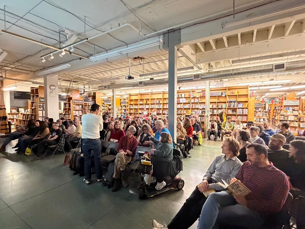 Crowd of people sitting with bookshelves in back. 