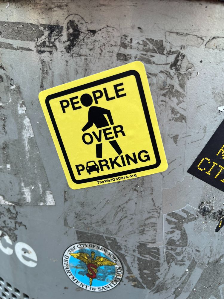 Square yellow sticker showing traffic sign image of a pedestrian with words "People Over Parking" on it. Stuck to the side of a grey garbage can.
