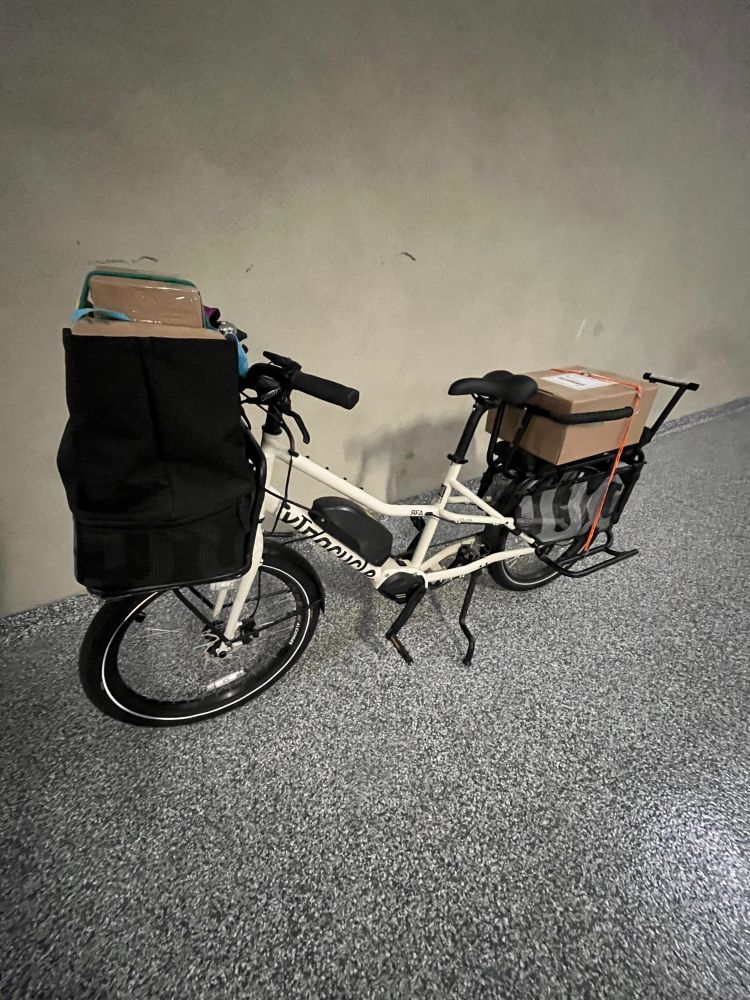 White Xtracycle cargo bike with black front carrier loaded with cardboard boxes and rear rack with cardboard box on it. Bike is in garage with grey/black speckled floor.
