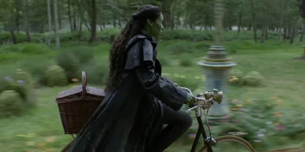 Still from Wicked showing Elphaba, wearing all black on a bike with a wicker basket on the back. In the background is green grass and trees and a lamp post.