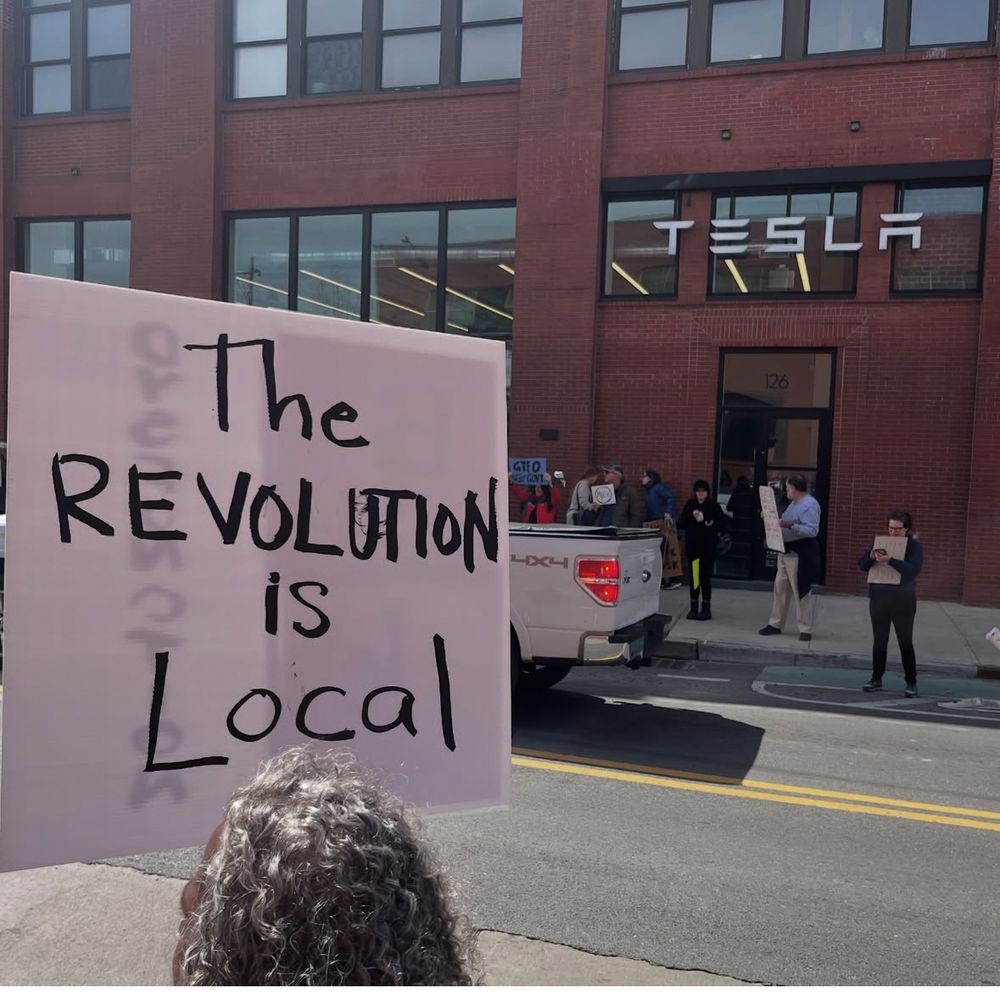 A protester holds a sign saying "The revolution is local" across the street from a Tesla dealership in Brooklyn, New York.