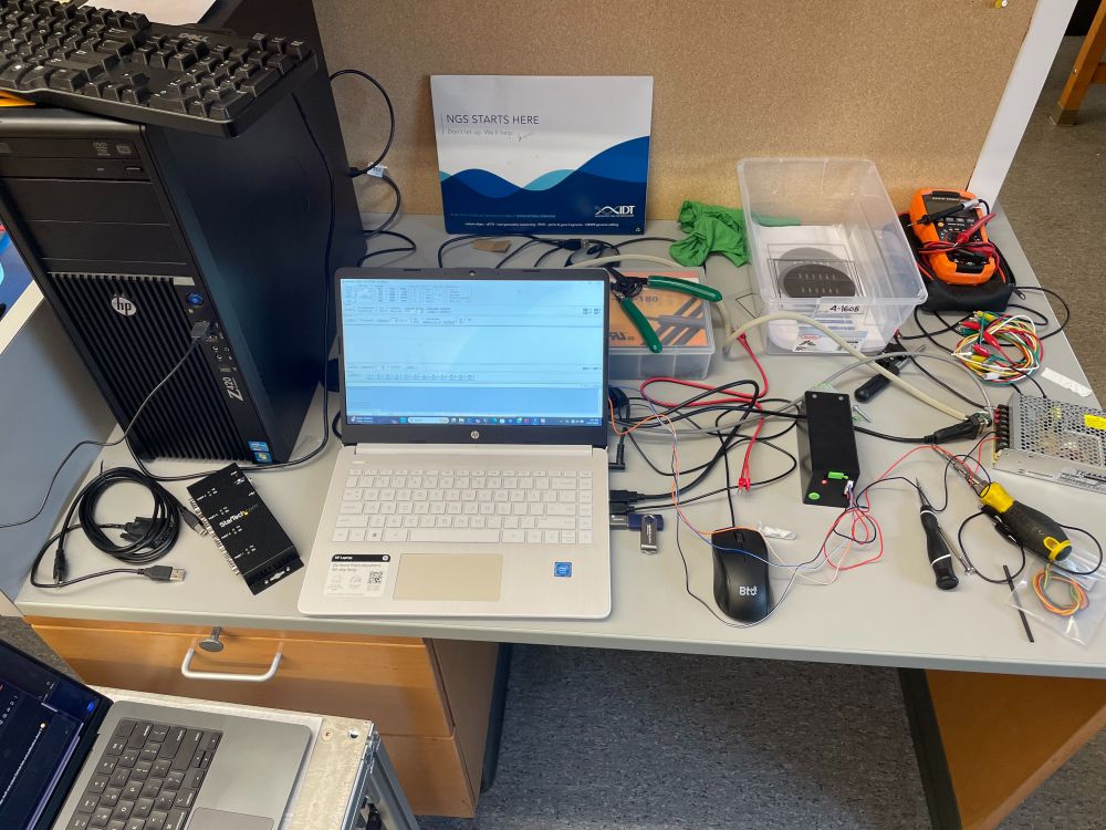 a right mess of a workstation with 3(?) computers, scattered tools and electronics equipment, some photomasks(?), some DNA(?), and a jackass selector valve
