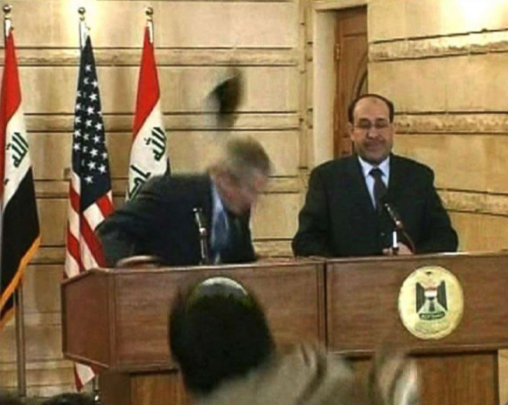Screenshot image from a televised speech capturing when George W Bush attempts to duck a flying shoe thrown at him.  
