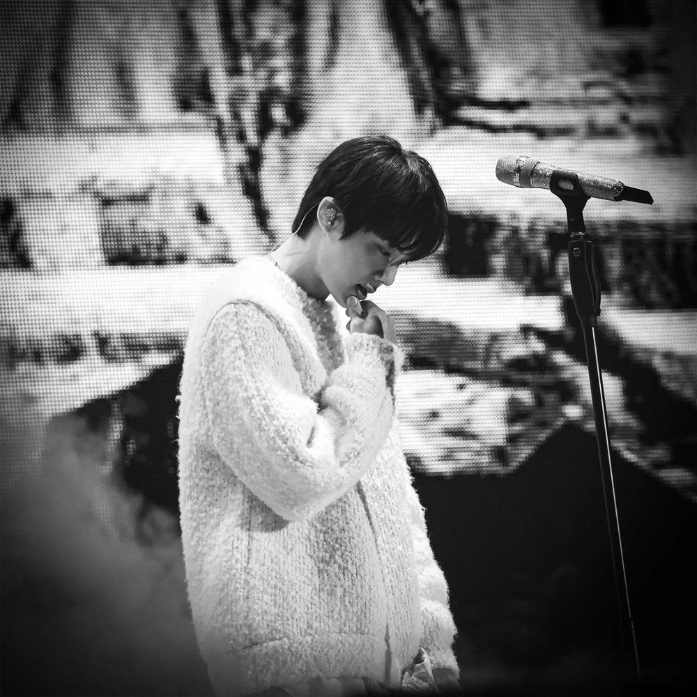 Member Jin of the korean band BTS , black and white image. He is standing with his head looking at the floor , a mic in a stand by his side and waterfalls in a screen as background