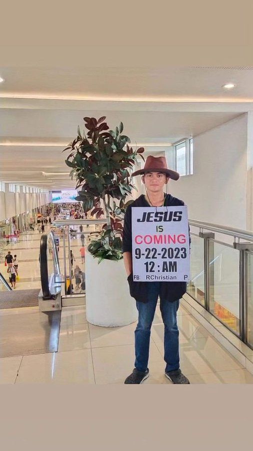 Weird person in a weird hat standing next to an escalator at an airport, maybe? wearing a big sign that says "Jesus is coming 9-22-2023 12:AM" 
Jesus didn't come then or today. Maybe next year. 