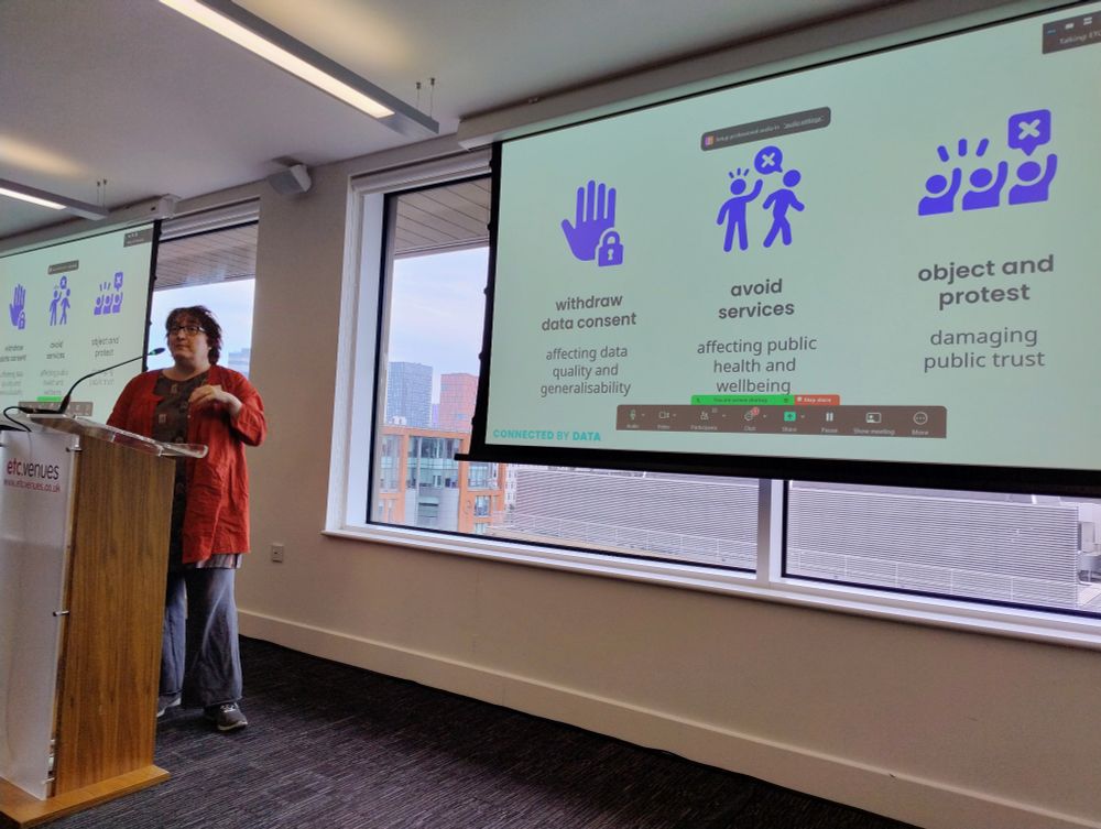 Jeni Tennison speaking at a conference; the PowerPoint slide in view highlights problems that arise if individuals don't engage/ trust govt data - they withdraw data consent, avoid services and object & protest