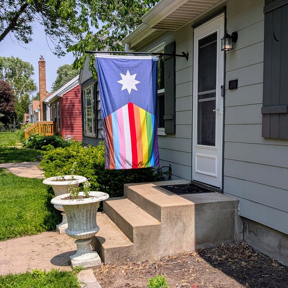 Picture of the MN state flag with pride rainbow stripes flying in front of a home.