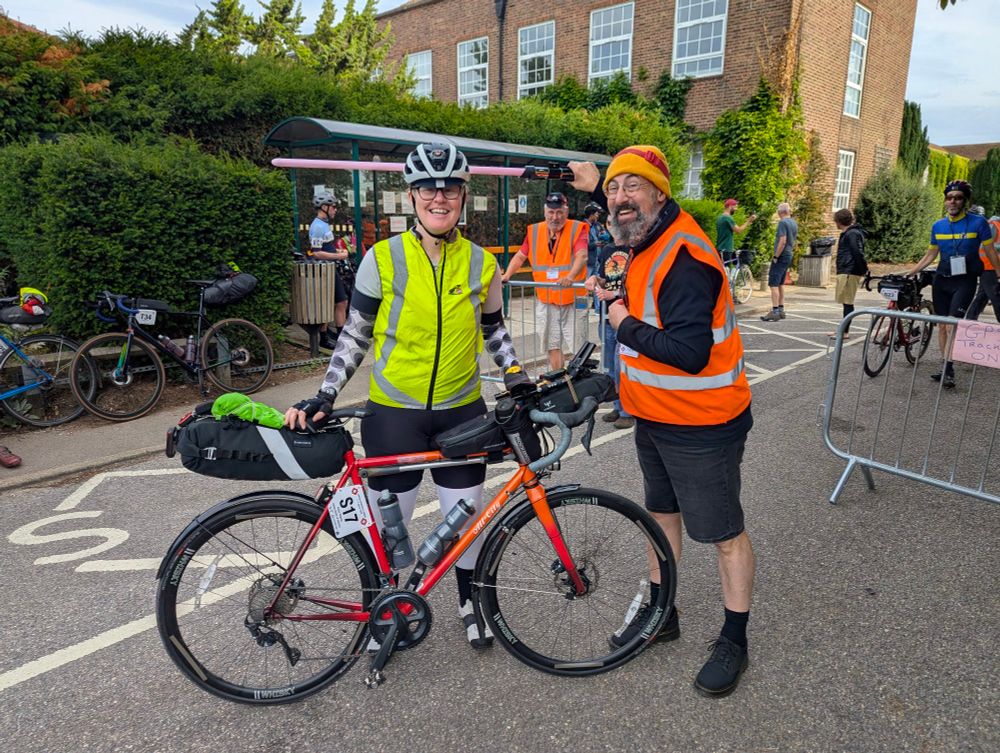 A woman in cycling kit wearing a yellow hi-vis vest stands behind her bike, loaded with multiple bags of gear, while a man in an orange hi-vis vest holds a pink light saber behind her head. 