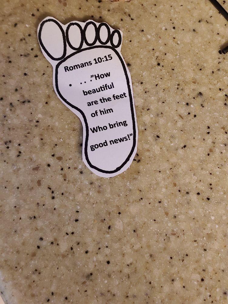 A small piece of paper cut into the shape of a foot on a counter. On the paper is a Bible verse Romans 10:15 ..."How beautiful are the feet of him Who bring good news!" Because my cousin is now in with God's foot fetish.