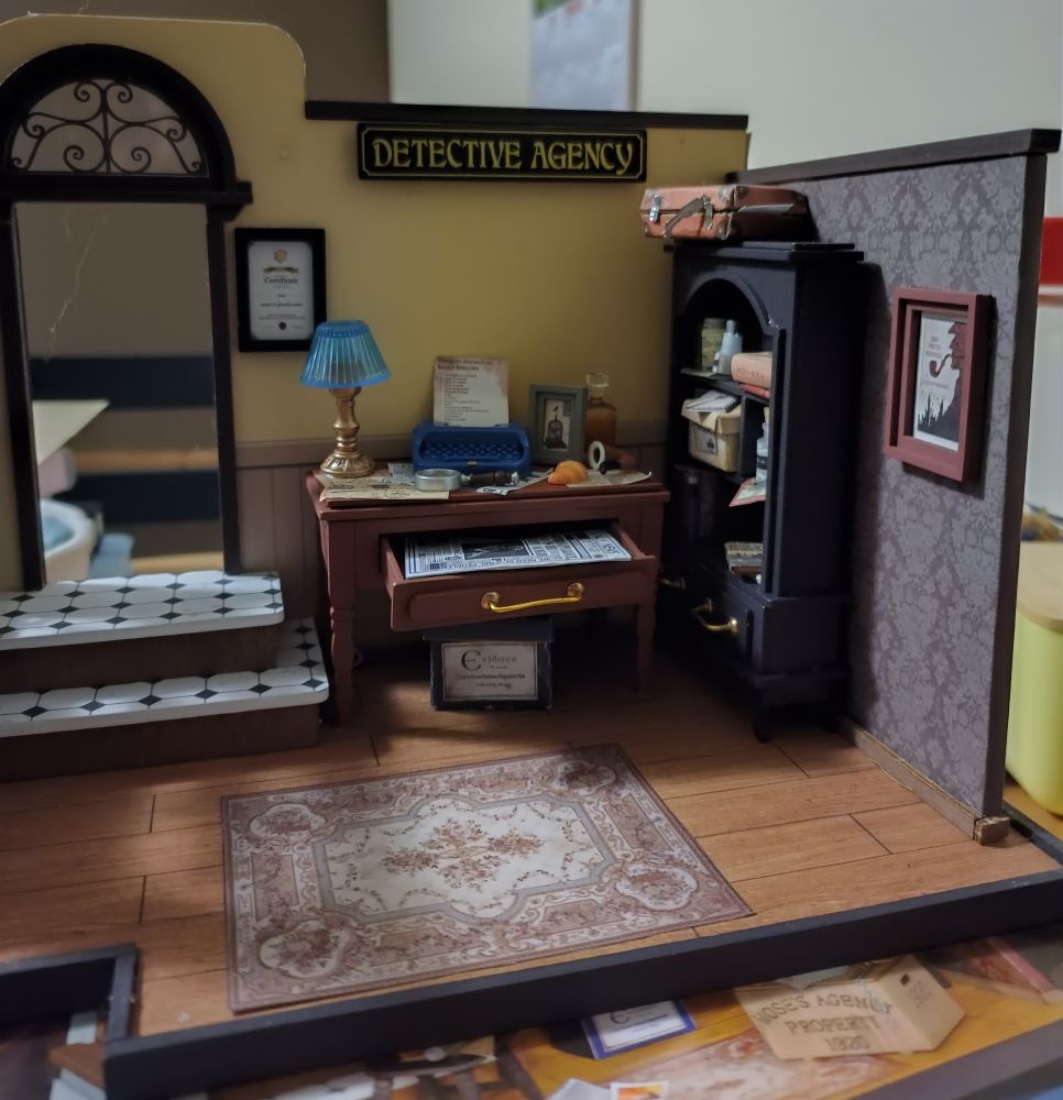 Miniatura a medio montar, que representa el gabinete de un detective tipo Sherlock Holmes, con papel pintado, un escritorio atestado y una estantería con libros, cajas y demás 
