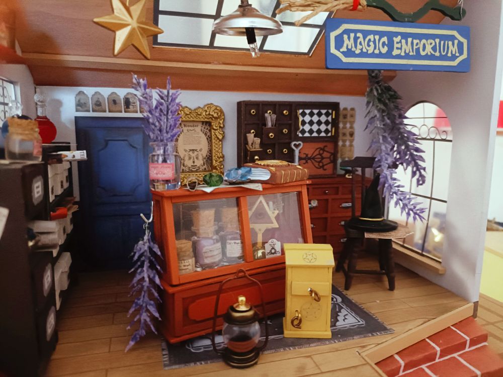 Un diorama de una tienda de magia, con lavanda colgando del techo; muebles llenos de frascos, pergaminos y piedras preciosas; sobre una silla está el sombrero de la bruja y la escoba cuelga del techo con el cartel del establecimientom desde este ángulo se aprecia una ventana con arco a la derecha y una estrella que cuelga del techo  