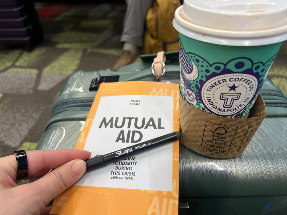 a copy of mutual aid by dean spade next to a cup of coffee on top of a sage colored carry on suitcase. mira’s hand is holding a pen in front of the book.