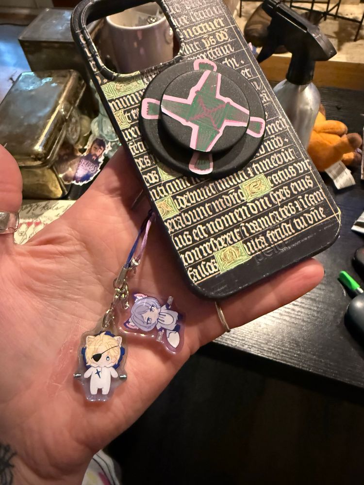 a phone case with imagery from the black book of hours at the morgan library and museum. the popsocket has a holographic cross of brigid sticker on it. two phone charms hang from it—one is fe3h dimitri in a lion suit and one is fe3h yuri in a wolf suit