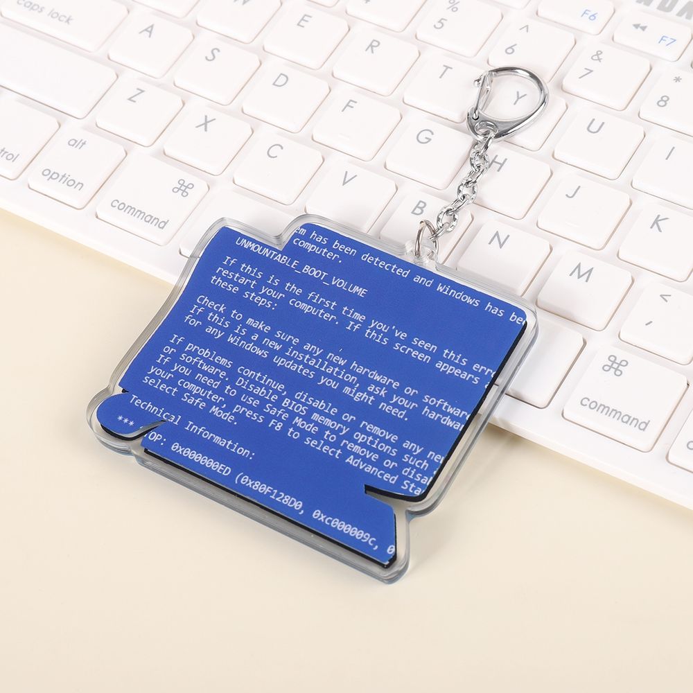 A photo of the back of the computer shaped keychain! It is the iconic Blue Screen of Death from Windows XP. It's misaligned on purpose, cutting off on the edges. The photo is taken on a desk near a keyboard.