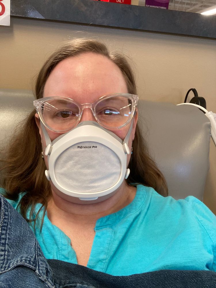 Selfie of me, a white lady with an enormous forehead, sitting in a blood donation chair, wearing a Flo mask respirator and safety glasses. 