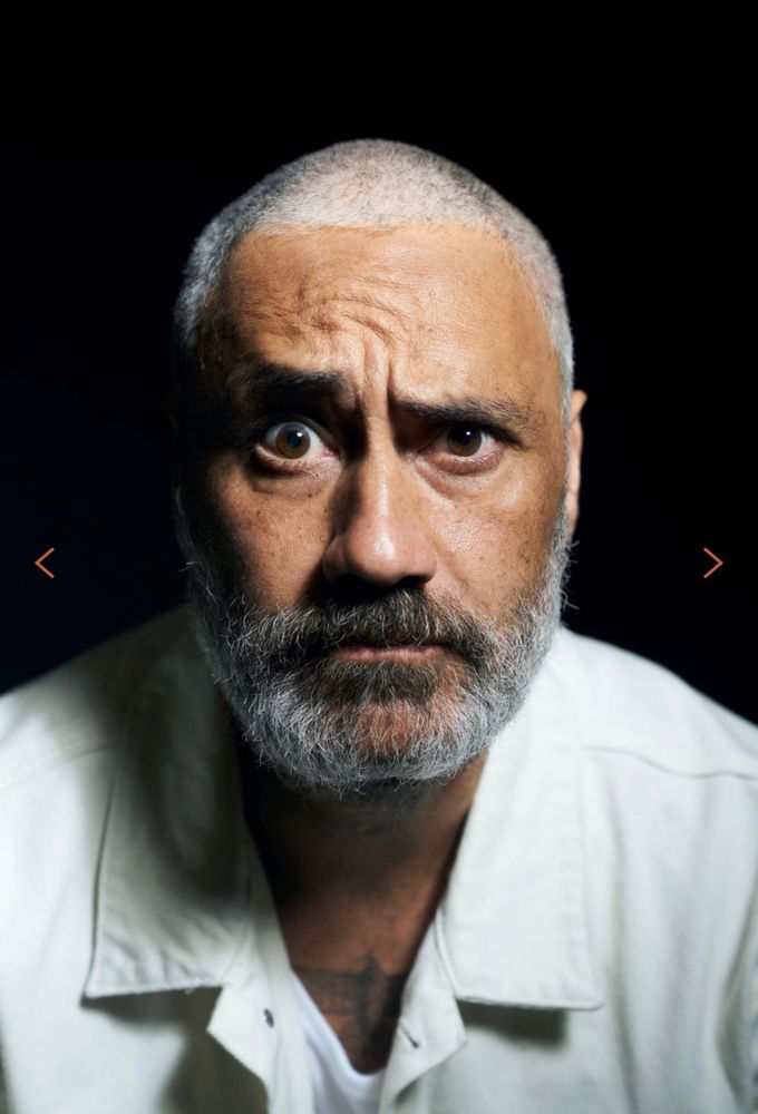 A close up photo of Taika Waititi. He’s wearing a white coat where part of his hawk tattoo is visible on his chest. He’s staring into the camera with a bewildered look. His head is shaved and he sports a beard. 