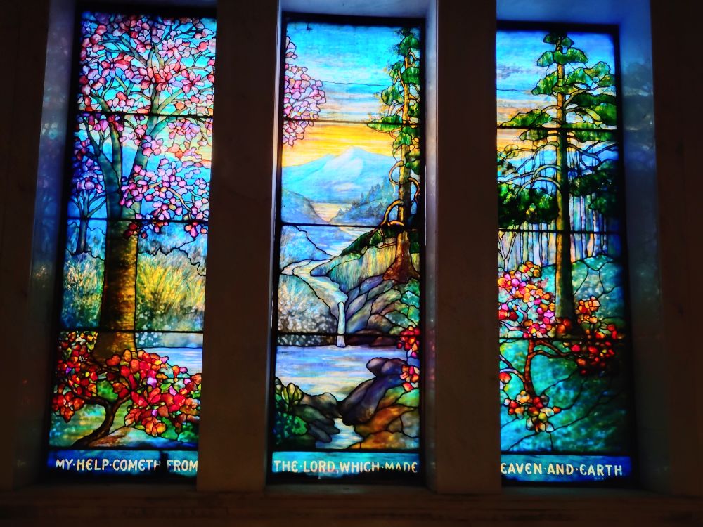 Three-panel Tiffany window showing a tranquil landscape. Verse at the bottom reads "My help cometh from the Lord which made Heaven and Earth."