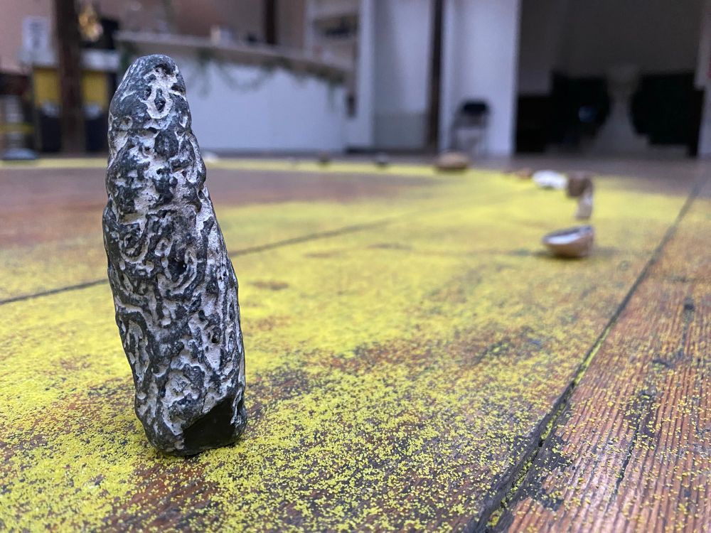A close up of a miniature standing stone, black and white patterned, stands on a wooden floor covered in yellow sand. In the background is a line of other stones, just out of focus. 