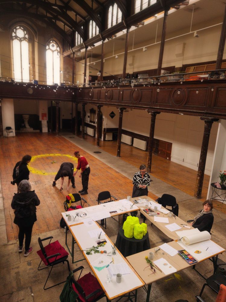 A group of people within an old church engaged in a series of tasks. Some are around a square table covered in paper and materials. Some are standing near a large yellow circle made of sand.