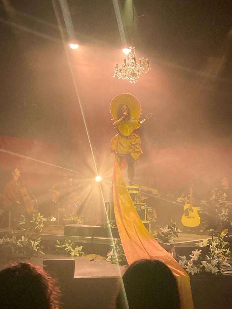 The finale of NO APOLOGIES, a brightly lit yellow stage full of musicians surrounding a performer standing on flight cases dressed as Kurt Cobain in a yellow gown that represents the sun. She is singing to the gathered audience. 