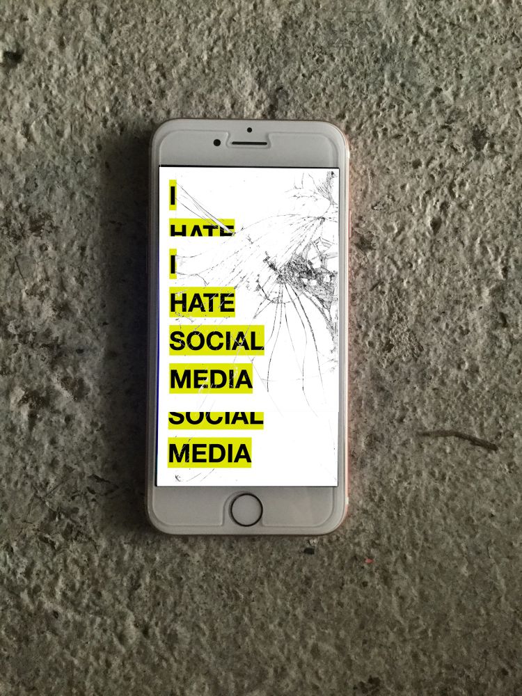 A smashed iPhone laying on a concrete floor. The screen is white and black and yellow hols text ‘I hate social media’ 