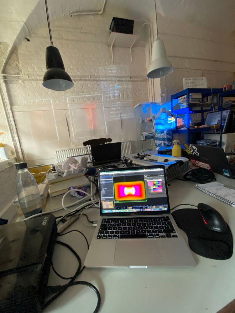 A laptop showing a bright orange and pink pattern sits on a white desk surrounded by cables and projectors. Projections cover the walls from different sources. 