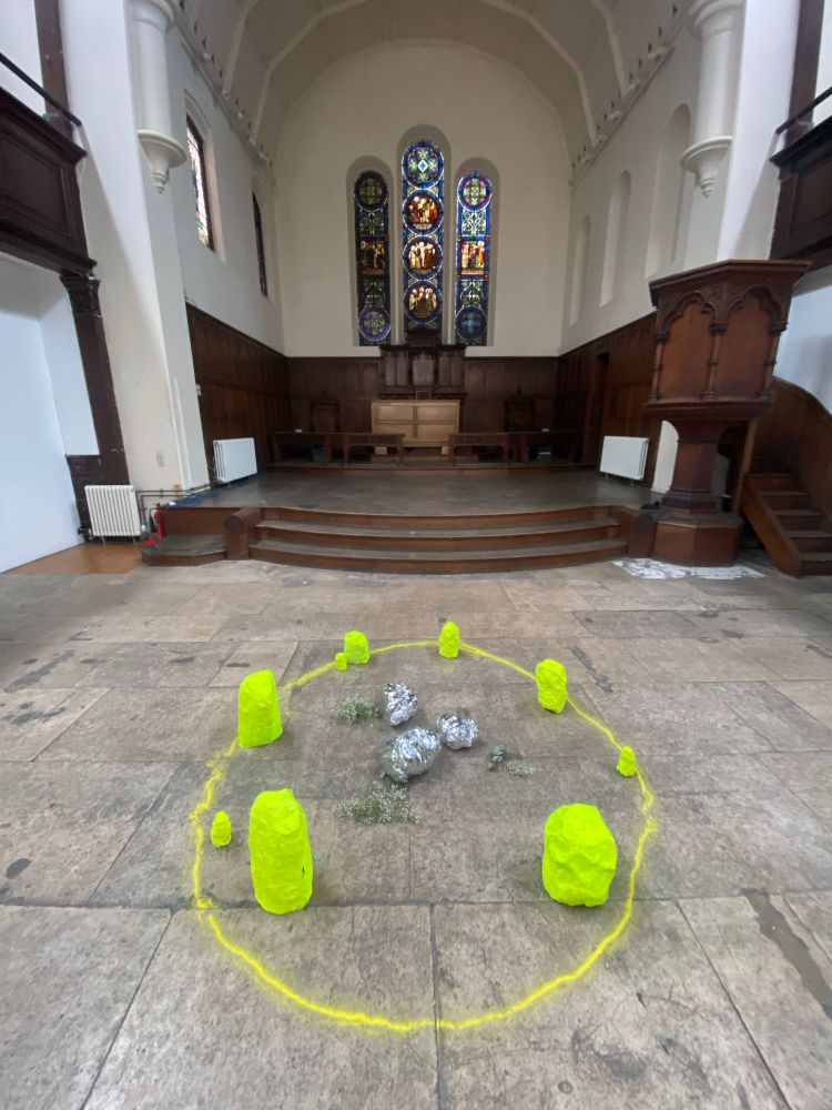 An old church with a grey concrete floor: in the centre of the floor is a stone circle made of yellow and silver paper stones, bright yellow sand, and flowers. 