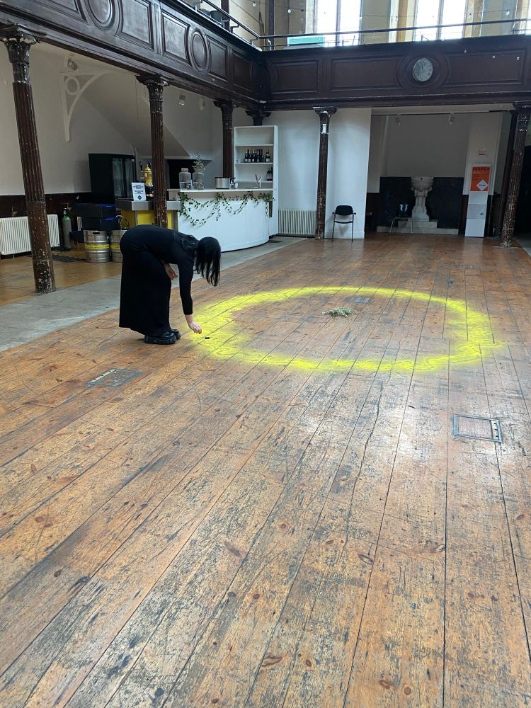 A person wearing a black skirt and top bends over placing a stone within a large yellow circle on a wooden floor.