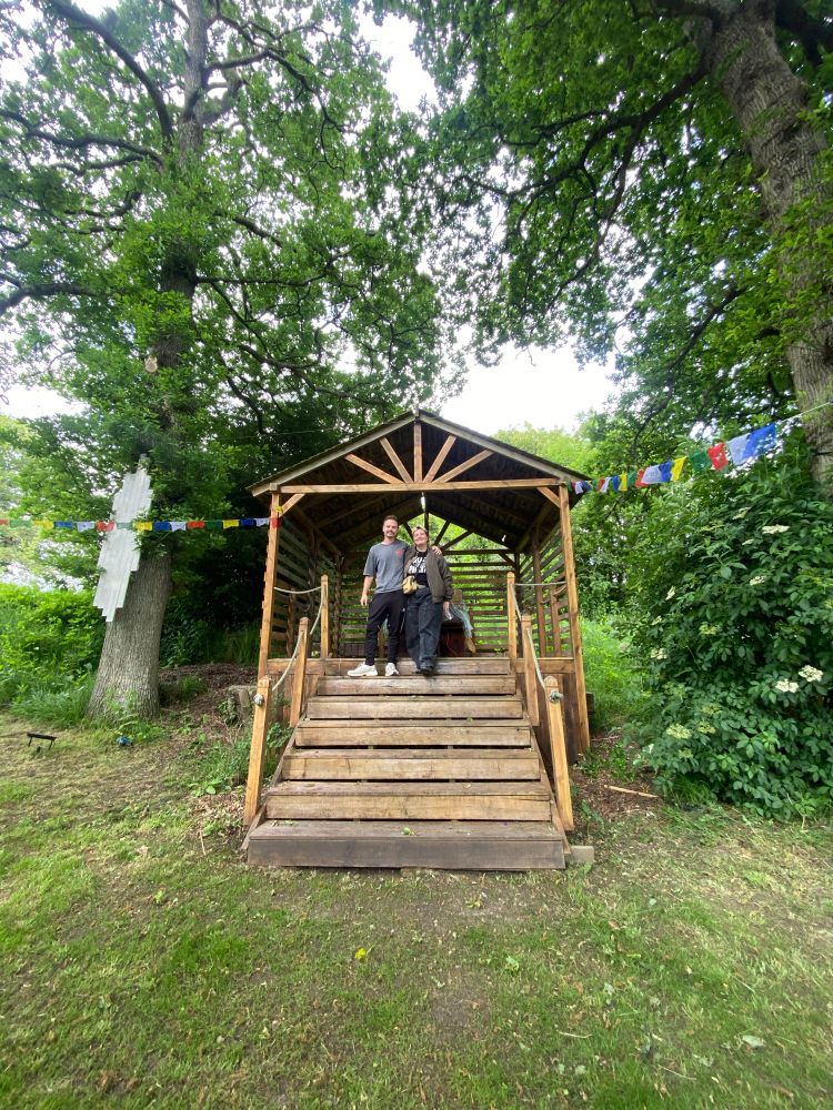 Two people stand on the steps of a small wooden stage in a woodland clearing. 
