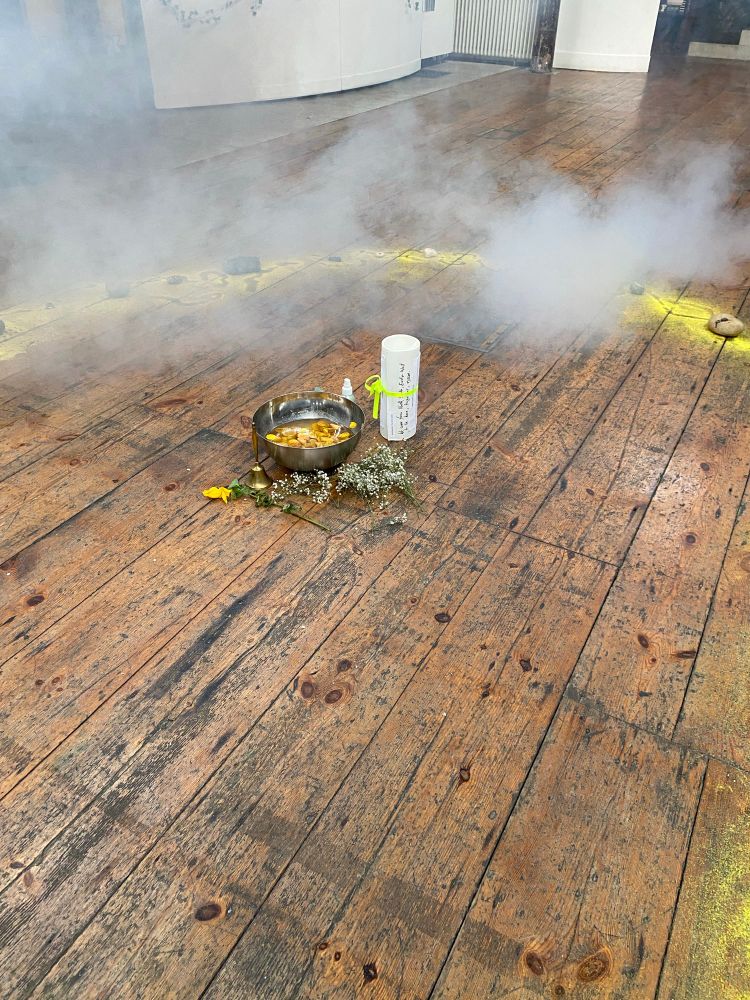 A ceremonial metal bowl in the centre of a yellow circle of sand filled with water and yellow rose petals. Next to it is a paper scroll tied with a yellow ribbon. Smoke drifts in from the top right of the image. 