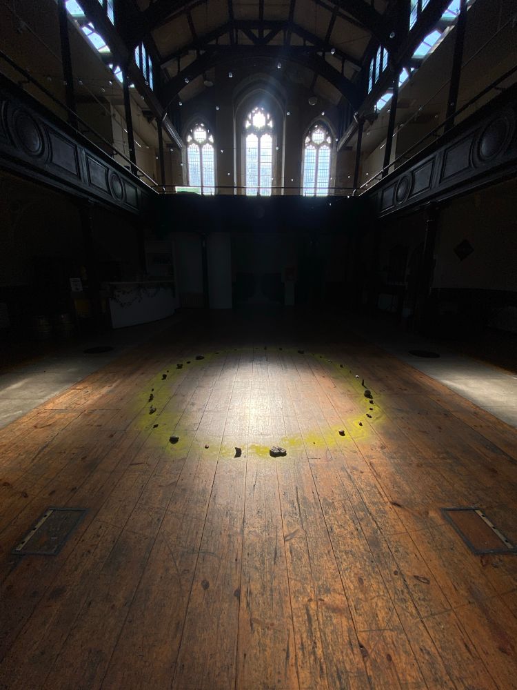 A large church hall, with soft morning light through tall windows illuminating a circle of stones on the wooden floor.