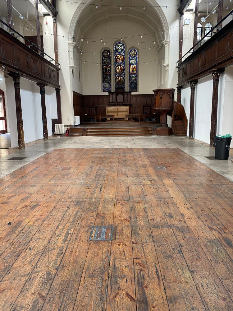 An empty church hall with a wooden floor and tall stained glass windows at one end 